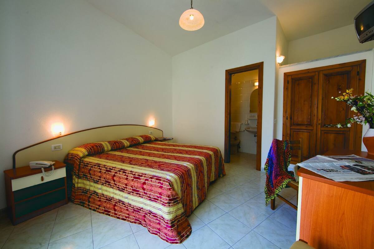 A bedroom with a bed, bedside table, lamp, and a wooden door leading to another room.