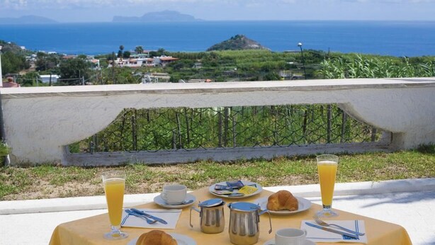 A table set for breakfast with a view of the ocean and a hill in the background. - Hotel al Bosco (Forio)