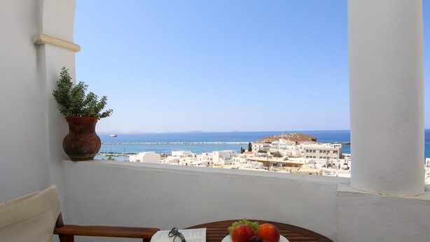 A balcony with a view of the sea and buildings, a wooden table with a book and fruit. - Panorama (Naxos)