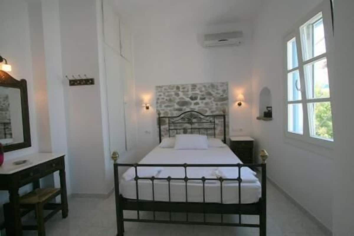 Ein Schlafzimmer mit einem Stein-Kopfteil, einem Bett mit weißer Bettwäsche, einem Nachttisch, einem Schreibtisch mit Stuhl und einem Fenster mit Vorhängen.
