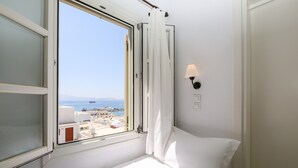 A hotel room with a bed, a window with curtains, a wall-mounted lamp, and a view of the sea and buildings. - Panorama (Naxos)