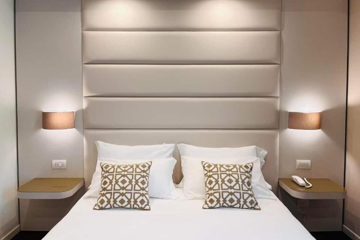 Een hotelkamer met een groot bed, twee bedlampjes, een nachtkastje met telefoon, en een hoofdeinde met horizontale panelen.