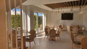 A spacious room with multiple round tables and wicker chairs, large windows offering a view of greenery, and a flat-screen TV mounted on the wall. - Hospedaria Frangaria (Faro)