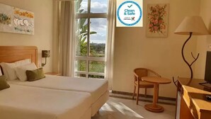A hotel room with two beds, a desk, a chair, and a TV. There is a window with a view of greenery and a sign indicating the room is clean and safe. - Hospedaria Frangaria (Faro)
