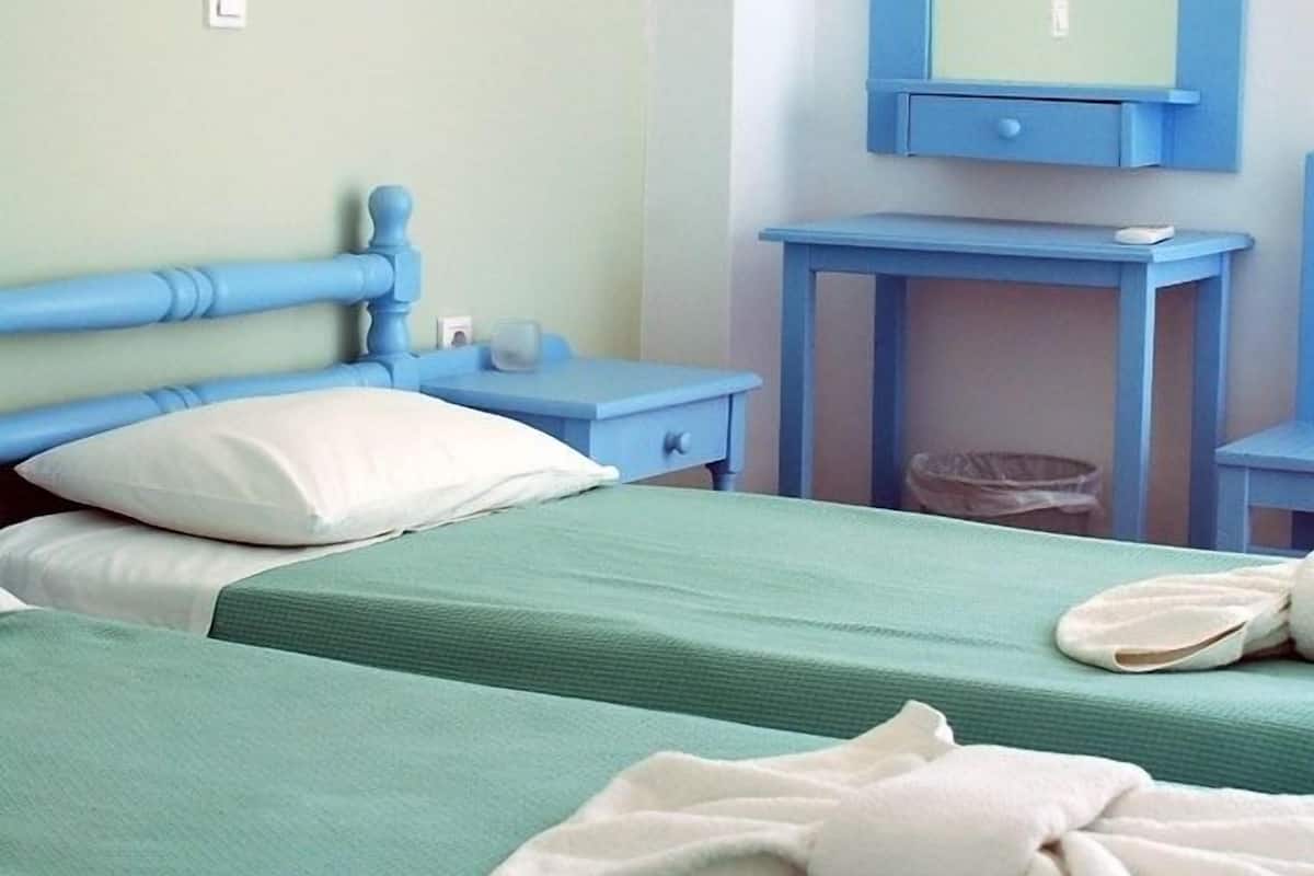 A room with two beds, each with a green bedspread and white pillows. There is a blue nightstand with a lamp, a blue dresser with a mirror, and a small trash bin.