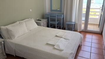 A neatly made bed with white linens and towels, a small bedside table with a remote control, a desk with a chair, a mirror, and a view of the sea through a sliding door.