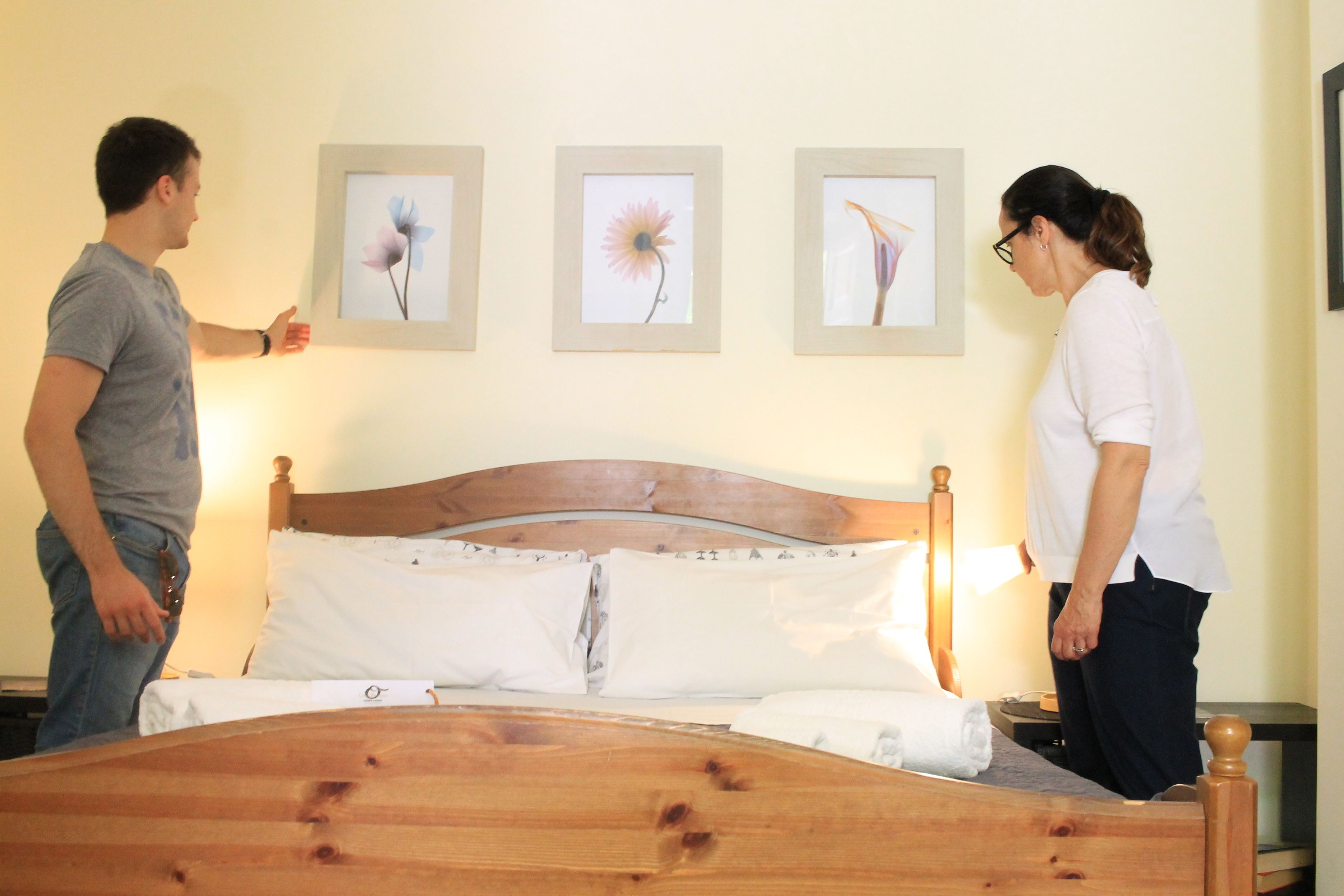 Un lit avec une tête de lit en bois, une literie blanche et trois tableaux représentant des motifs floraux, encadrés et accrochés au mur.