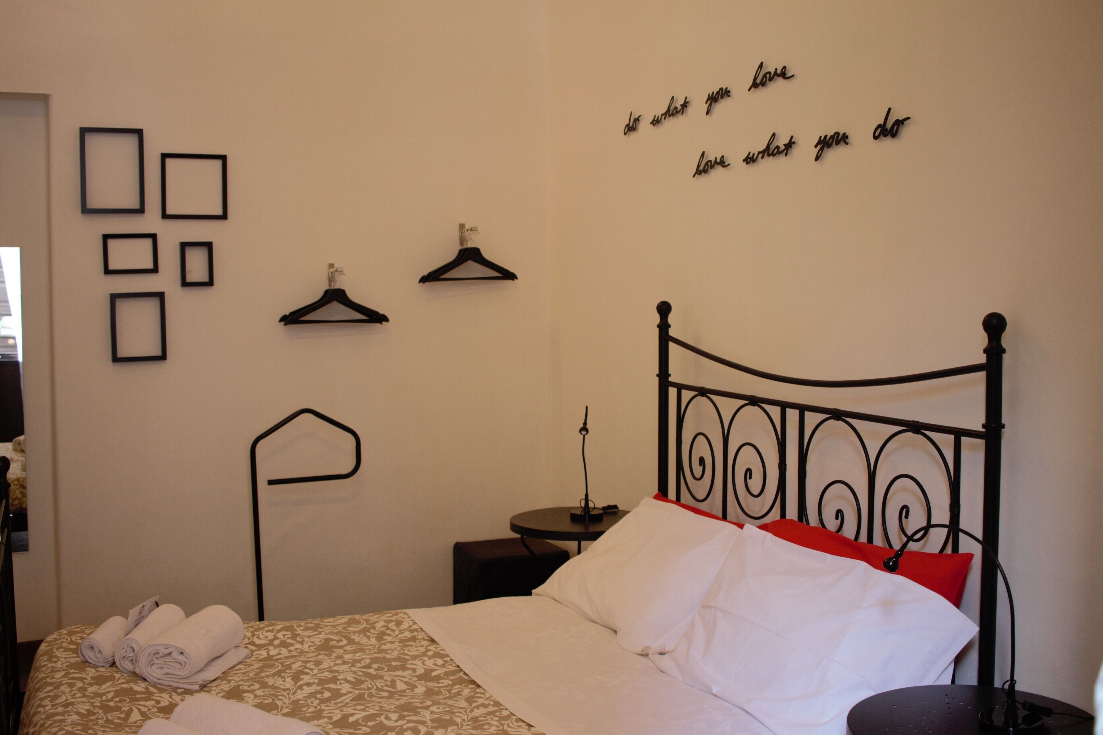 Una cama bien hecha con almohadas blancas y rojas, una mesita de noche con lámpara, y una pared con cuadros y una cita motivacional.