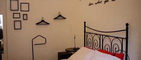 Una cama bien hecha con almohadas blancas y rojas, una mesita de noche con lámpara, y una pared con cuadros y una cita motivacional.