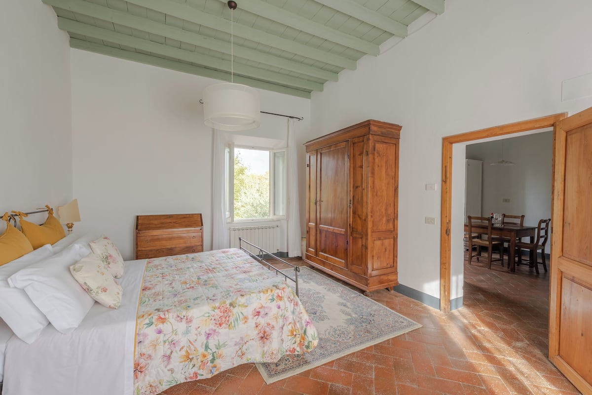 Un dormitorio con cama, armario de madera y una zona de comedor visible a través de una puerta abierta.