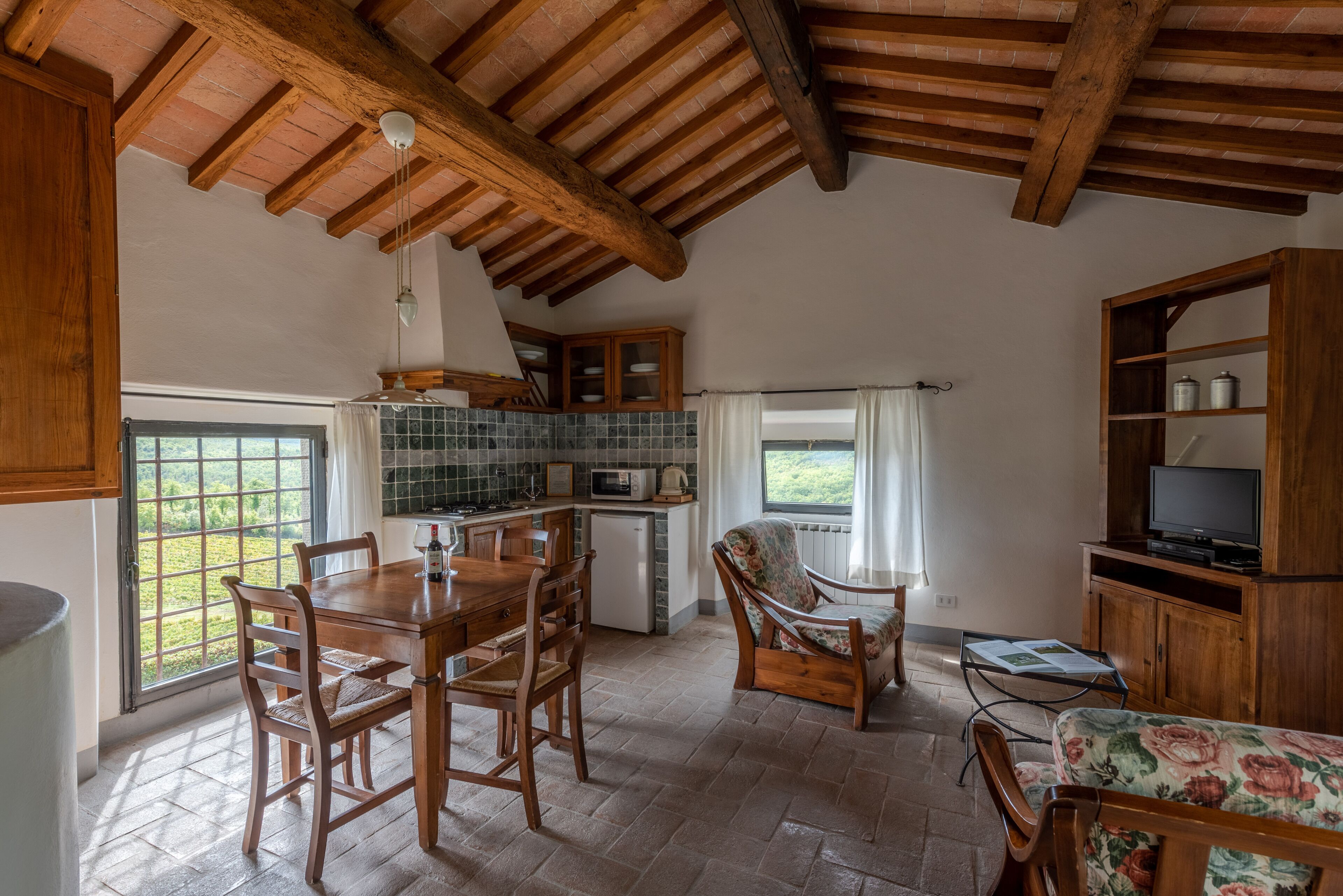 Una cocina amplia con vigas de madera, una zona de comedor con sillas, un televisor y vistas a zonas verdes exteriores.