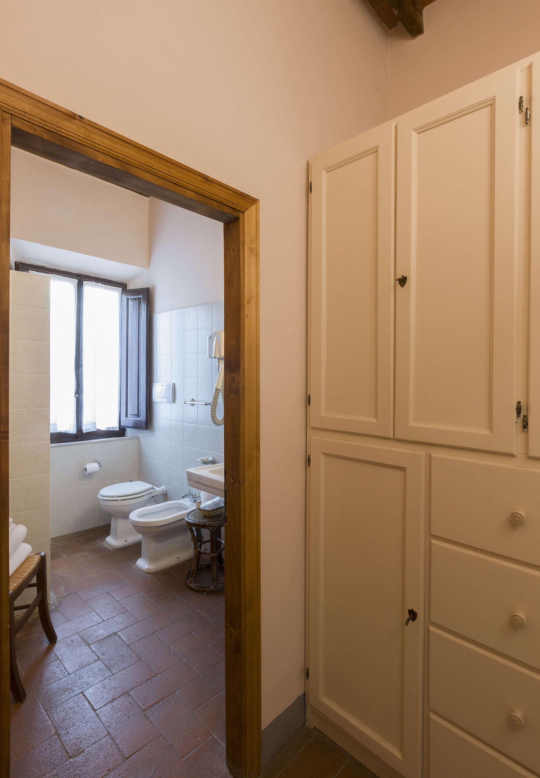 A bathroom with a toilet, bidet, and sink. There is a wooden door frame and a window with blinds.