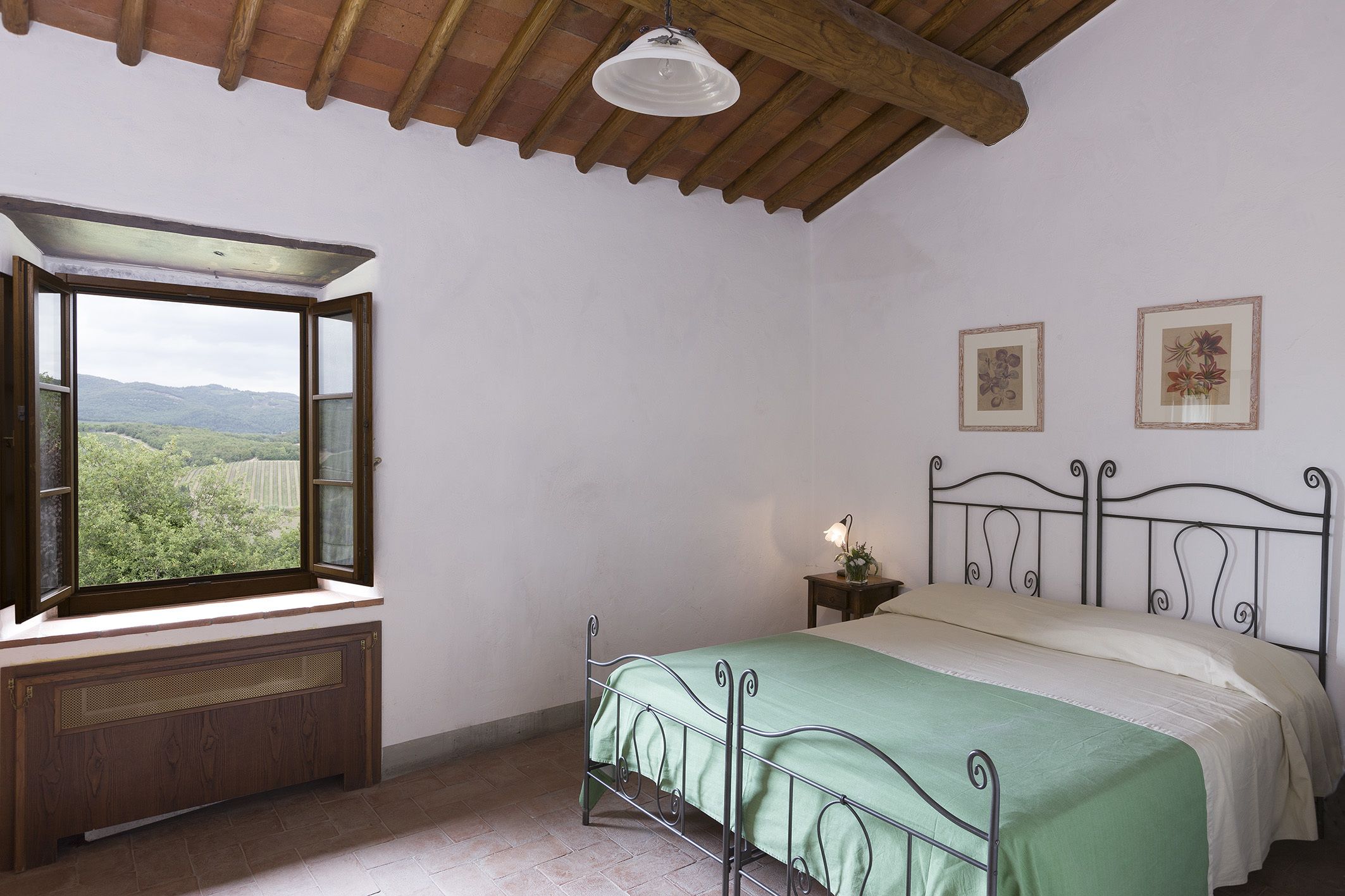 A bedroom with a bed, a window with a view of greenery, a radiator, and two framed pictures on the wall.