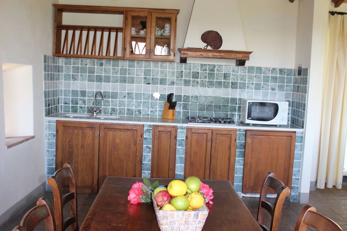 Una mesa de madera con sillas, una cesta de frutas y una cocina con microondas, fregadero y armarios.