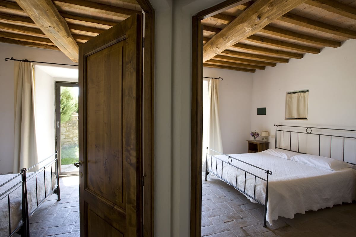 Un dormitorio con cama, vigas de techo de madera, una ventana con cortinas y una puerta que conduce a otra habitación.