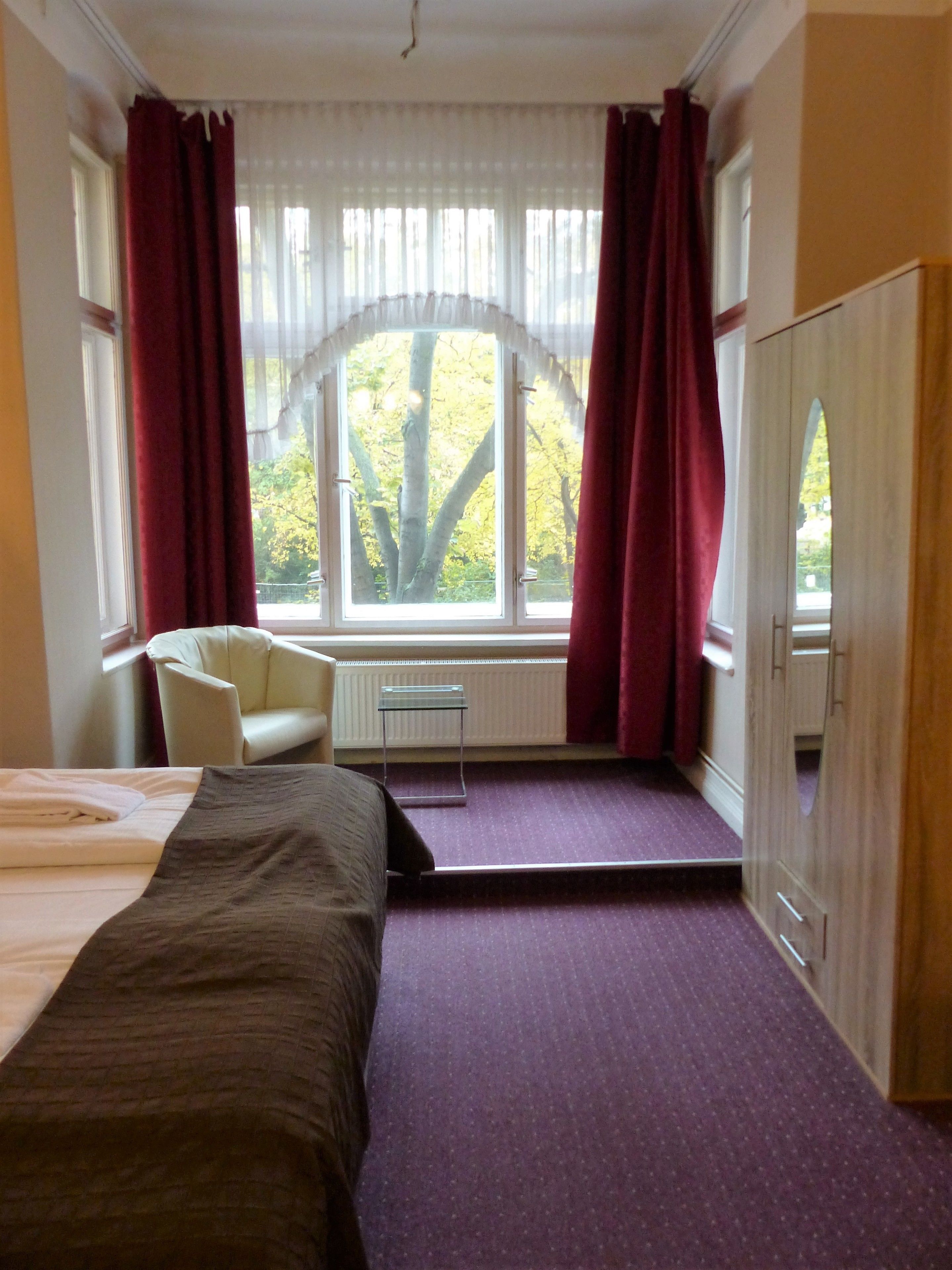 A hotel room with a bed, a chair, a small table, and a window with curtains.