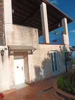 Um edifício com fachada de concreto, uma varanda de metal com guarda-corpo preto e uma janela pequena com persianas azuis.
