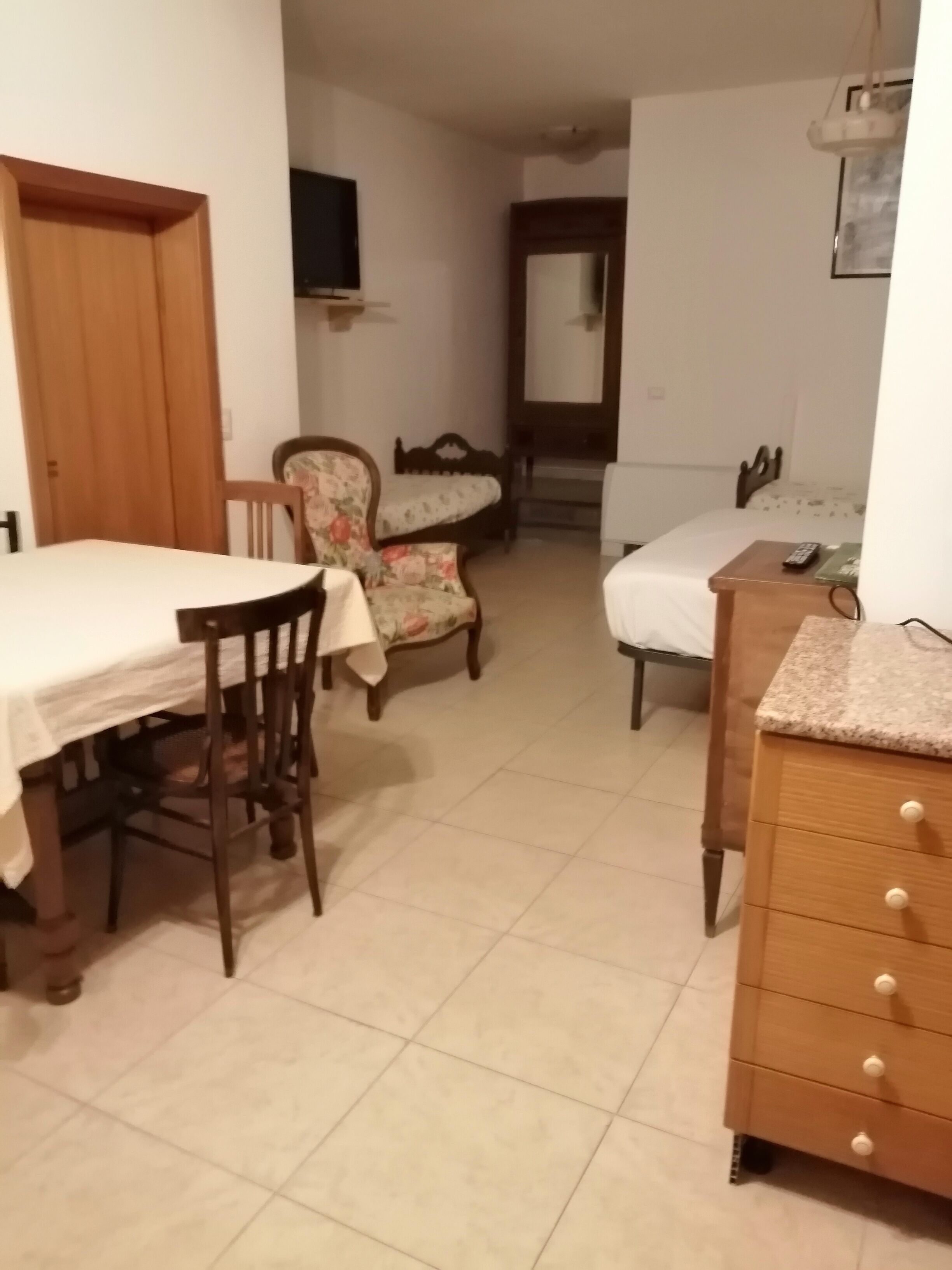 A compact room with a dining table, a floral armchair, a bed with a wooden headboard, a small dresser, and a mirror on the wall.