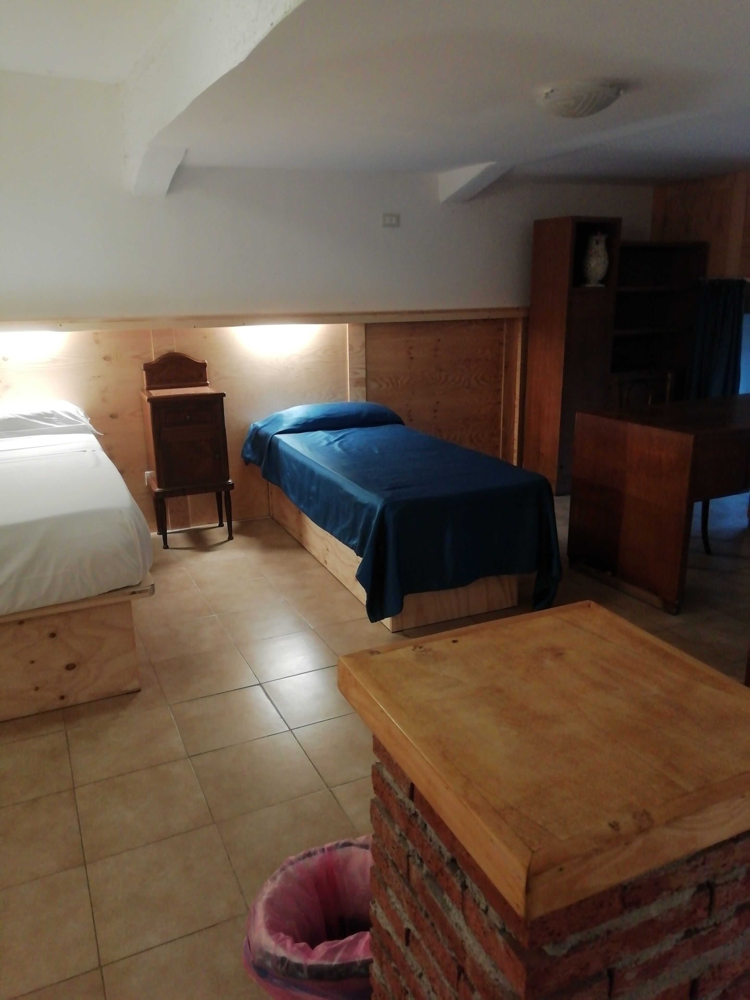 A dormitory room with two beds, a wooden desk, a small cabinet, and a trash bin.