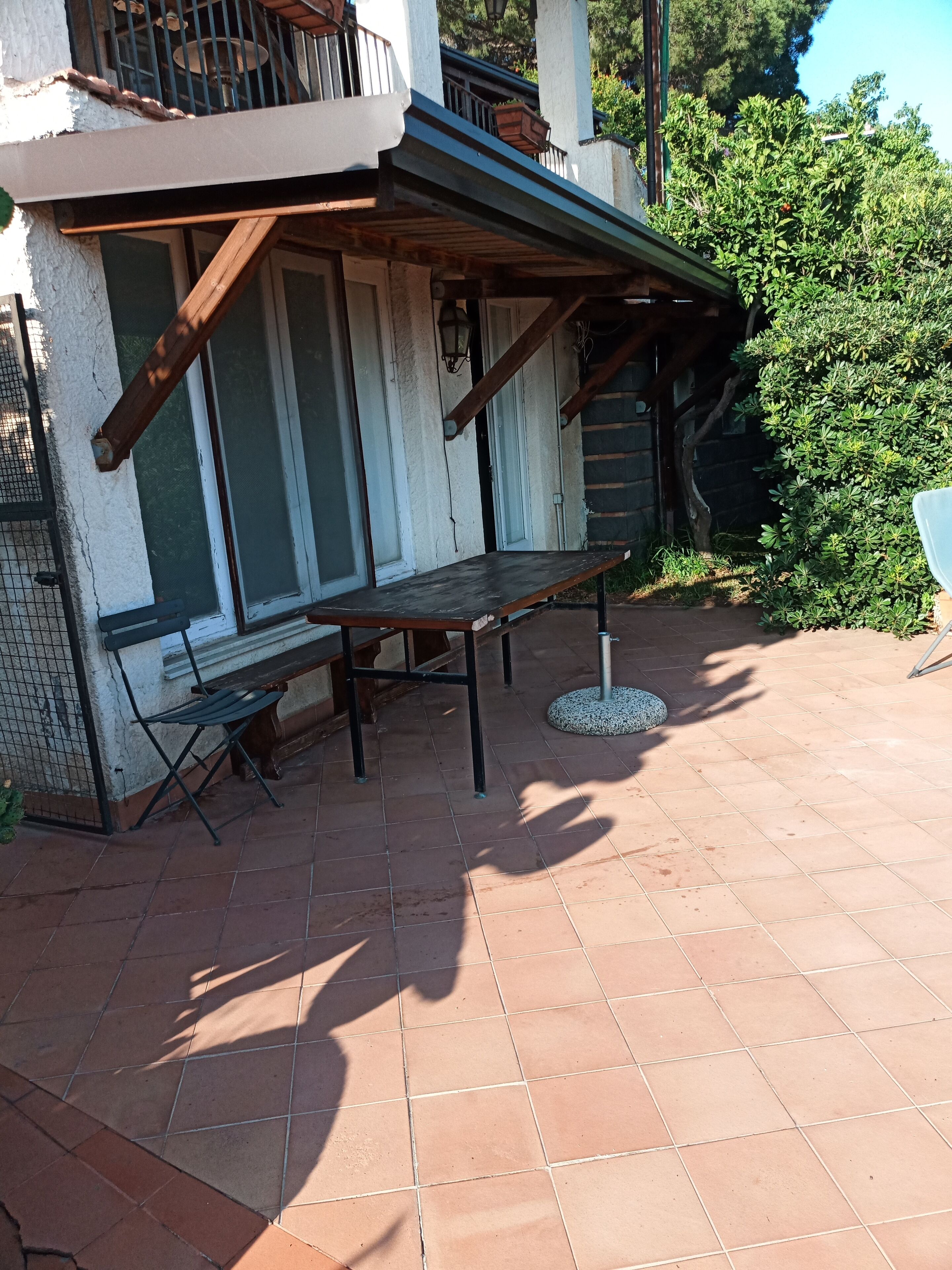 Eine Terrasse im Freien mit Holztisch und -stühlen, einem gefliesten Boden und Blick auf ein Gebäude mit Balkon.