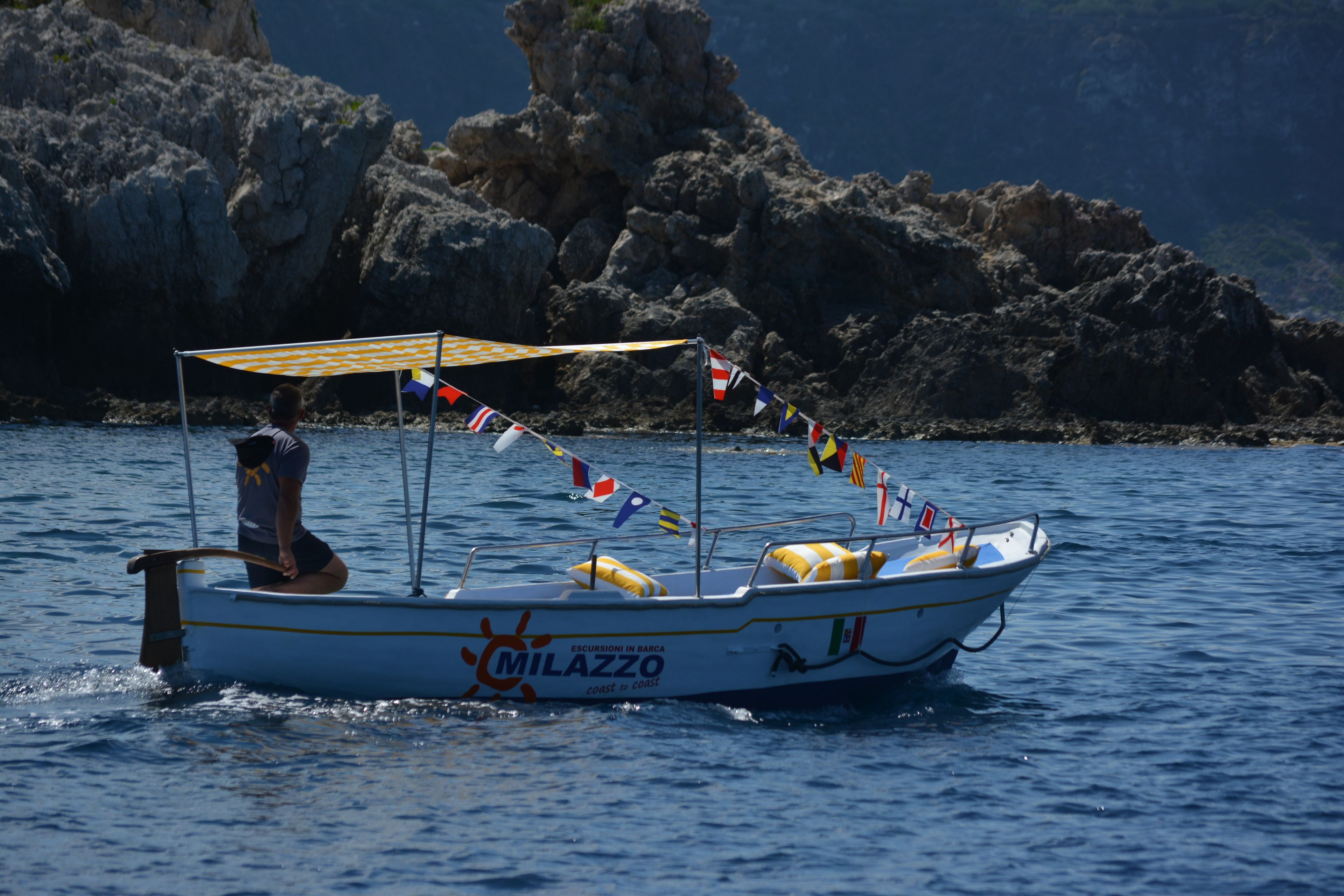 A boat named "Milazzo" with a canopy and colorful flags, sailing on water with rocky cliffs in the background.