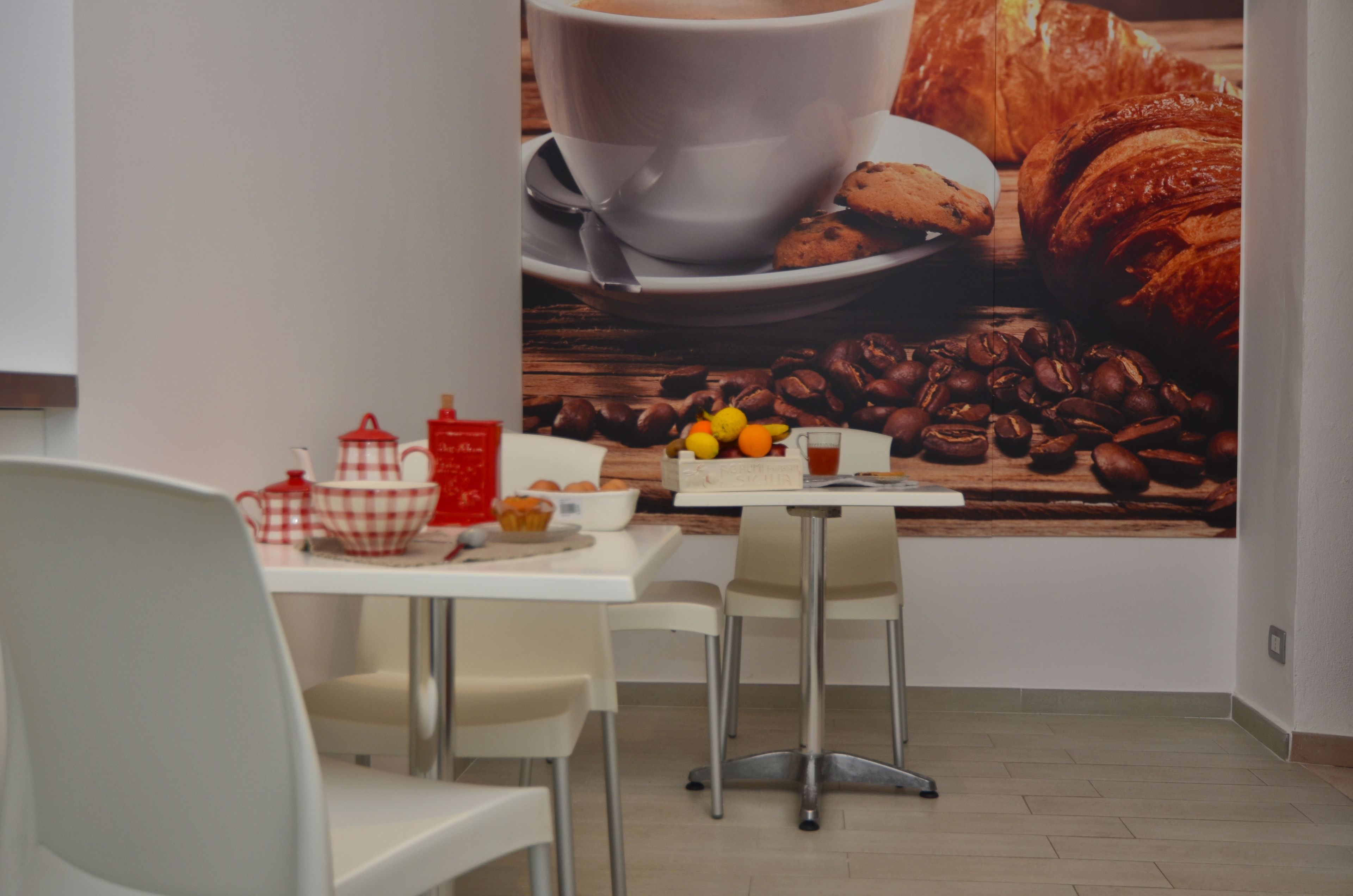 Een eetgedeelte met een tafel gedekt voor twee personen, met een witte tafelkleed, twee witte stoelen, een rood-wit geruit tafelkleed, een rode theepot, een witte beker, een witte kom, en een bord met koekjes en een croissant.