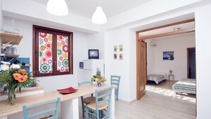 Ein Essbereich mit einem Holztisch, Stühlen und einer Blumenvase. Ein Fernseher steht auf einem Schrank und ein Fenster zeigt bunte Muster.