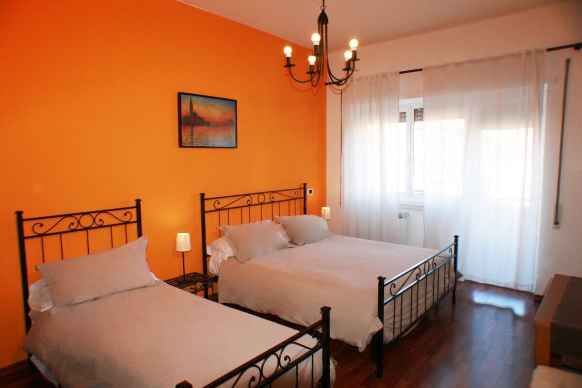 A room with two beds, a painting on the wall, a chandelier, and a window with curtains.