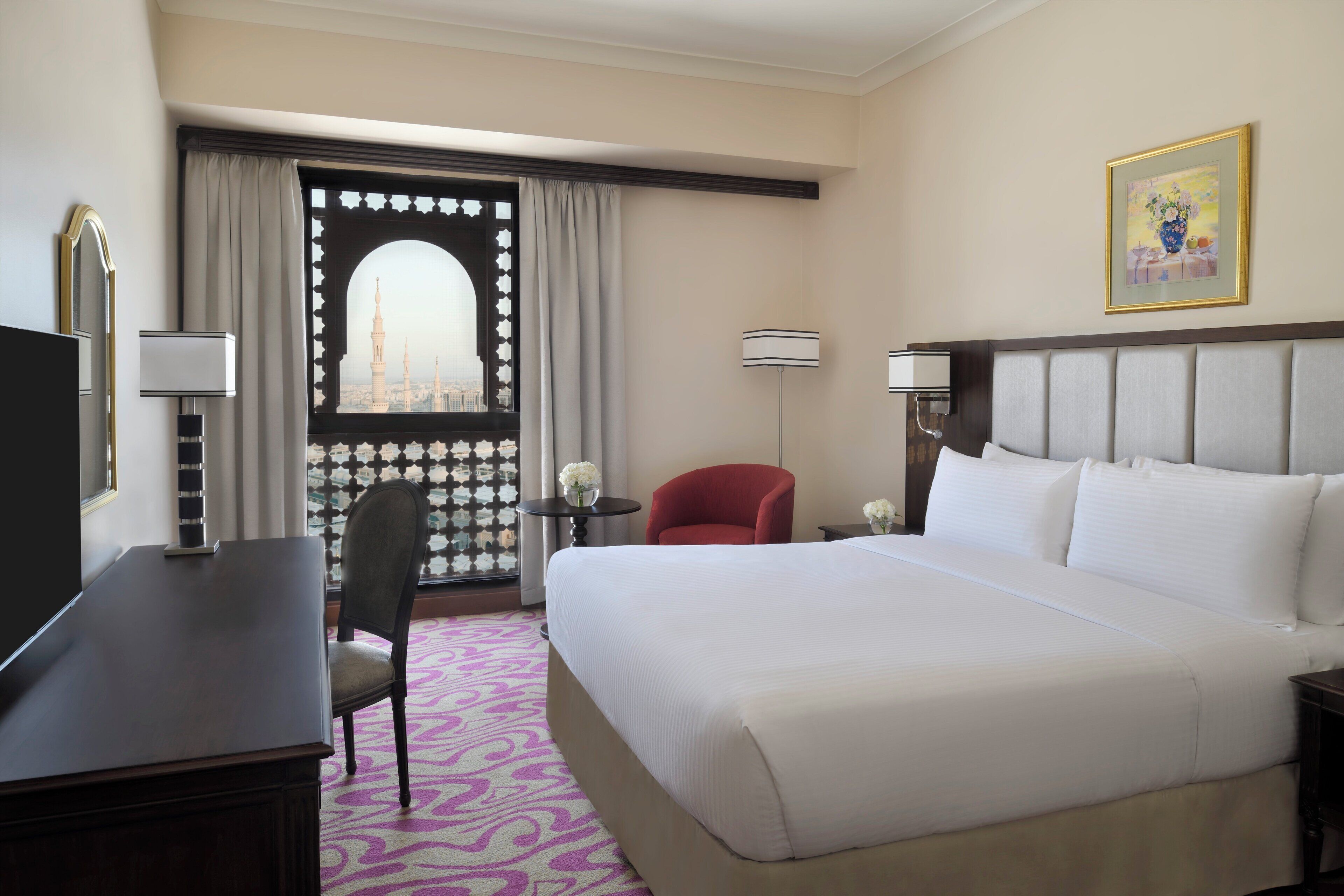 A hotel room with a large bed, a desk, a chair, a small table with a vase of flowers, a lamp, and a view of a cityscape through a window with a decorative screen.