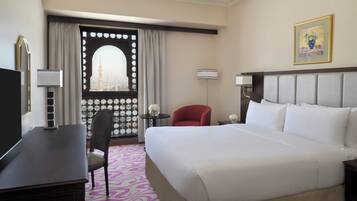 A hotel room with a large bed, a desk, a chair, a small table with a vase of flowers, a lamp, and a view of a cityscape through a window with a decorative screen.
