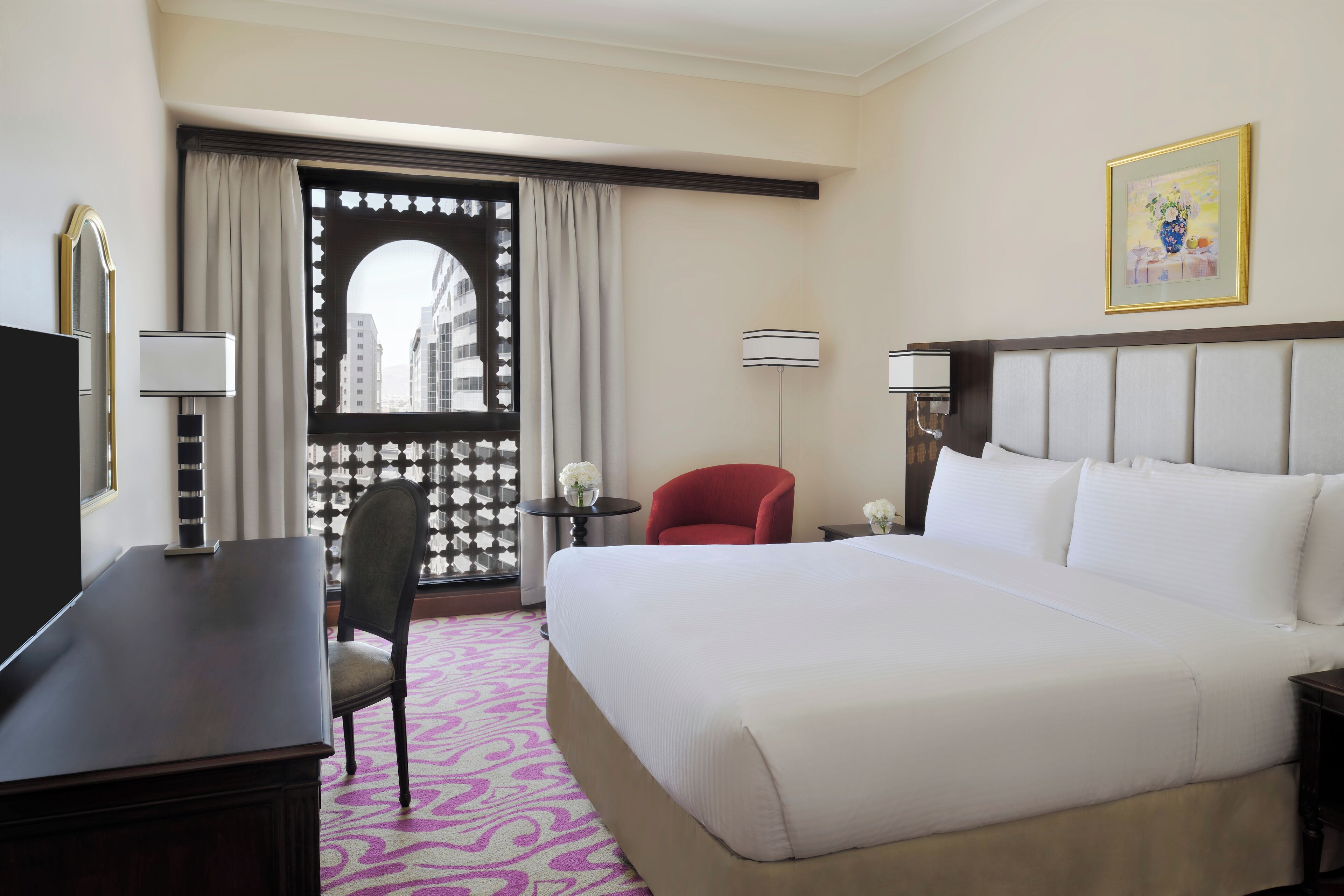 Une chambre d’hôtel avec un grand lit, un bureau, une chaise, une lampe et une vue sur l’extérieur, visible à travers une fenêtre avec des rideaux.