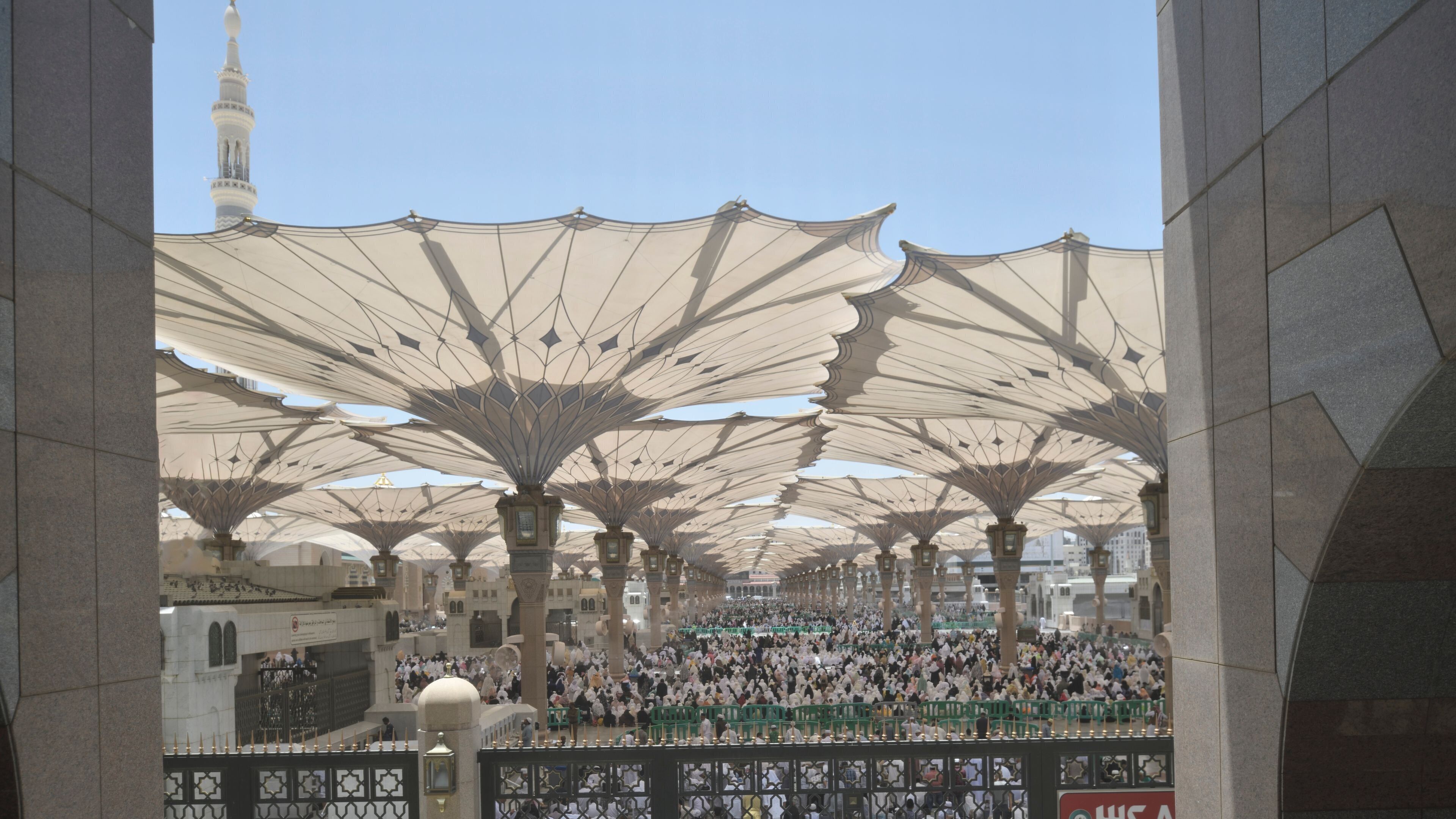A large covered area with a crowd of people, featuring a prominent minaret in the background.