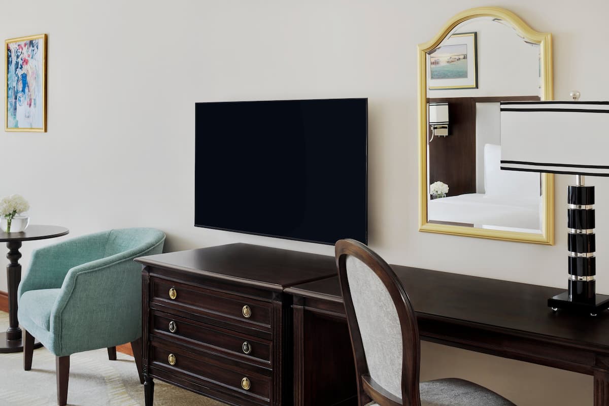Ein Hotelzimmer mit einem dunklen Holzbüro, einem blaugrünen Sessel, einem großen Spiegel und einem Flachbildfernseher.