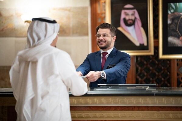 A man in a white robe handing over a card to a man in a blue suit at a reception desk with framed portraits in the background. - Intercontinental Madinah - Dar Al Iman by IHG (Madinah)