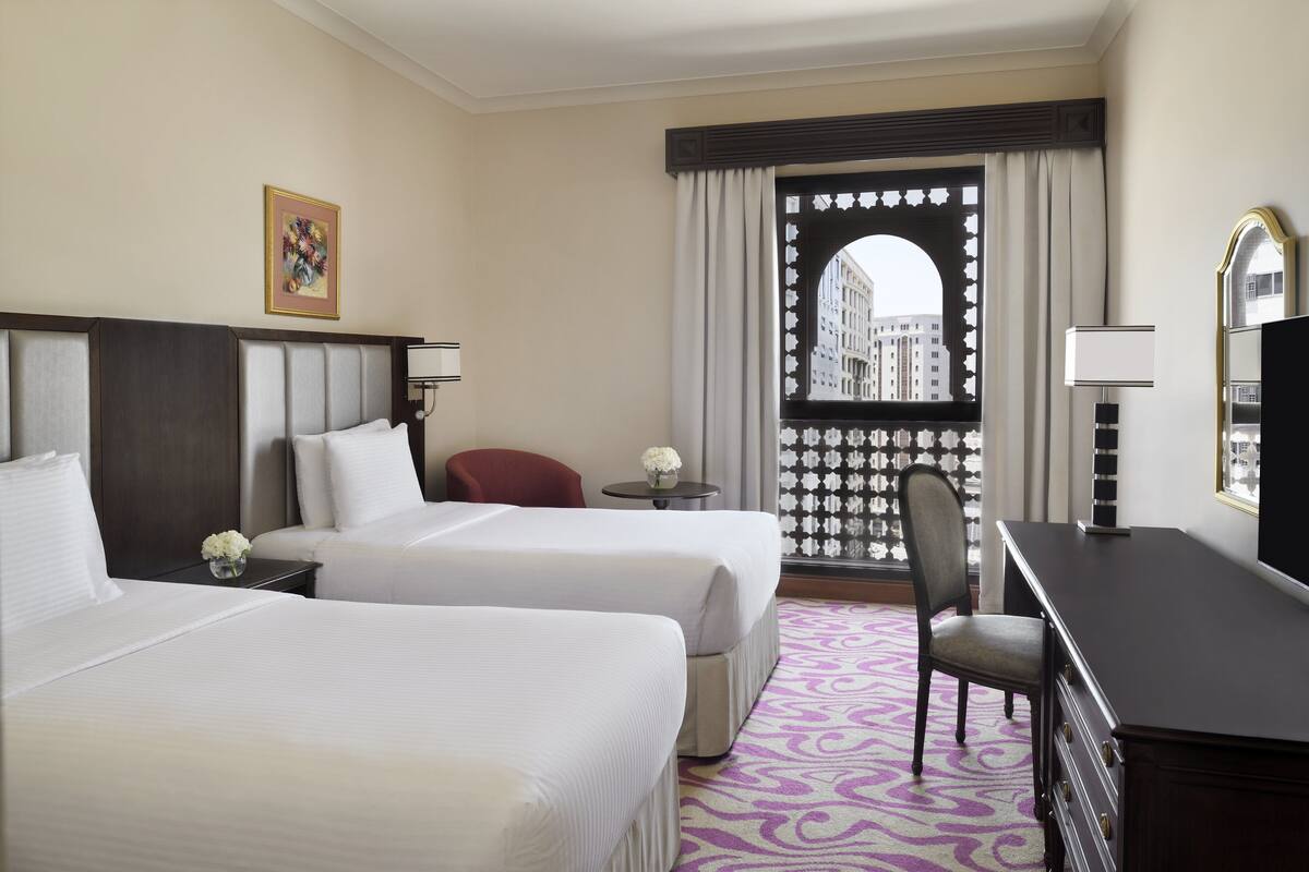 Ein Hotelzimmer mit zwei Betten, einem Schreibtisch, einem Stuhl, einem kleinen Tisch mit einer Vase sowie einem Fenster mit Blick auf Gebäude.