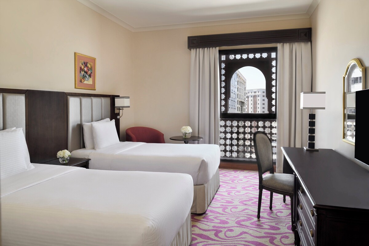 A hotel room with two beds, a desk, a chair, a small table with a vase, and a window with a view of buildings.