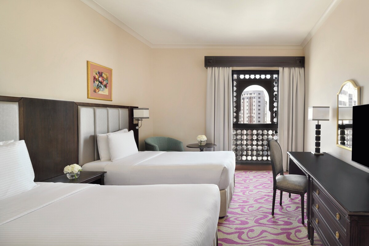 A hotel room with two beds, a desk, a chair, a small table with a vase, and a window with curtains.