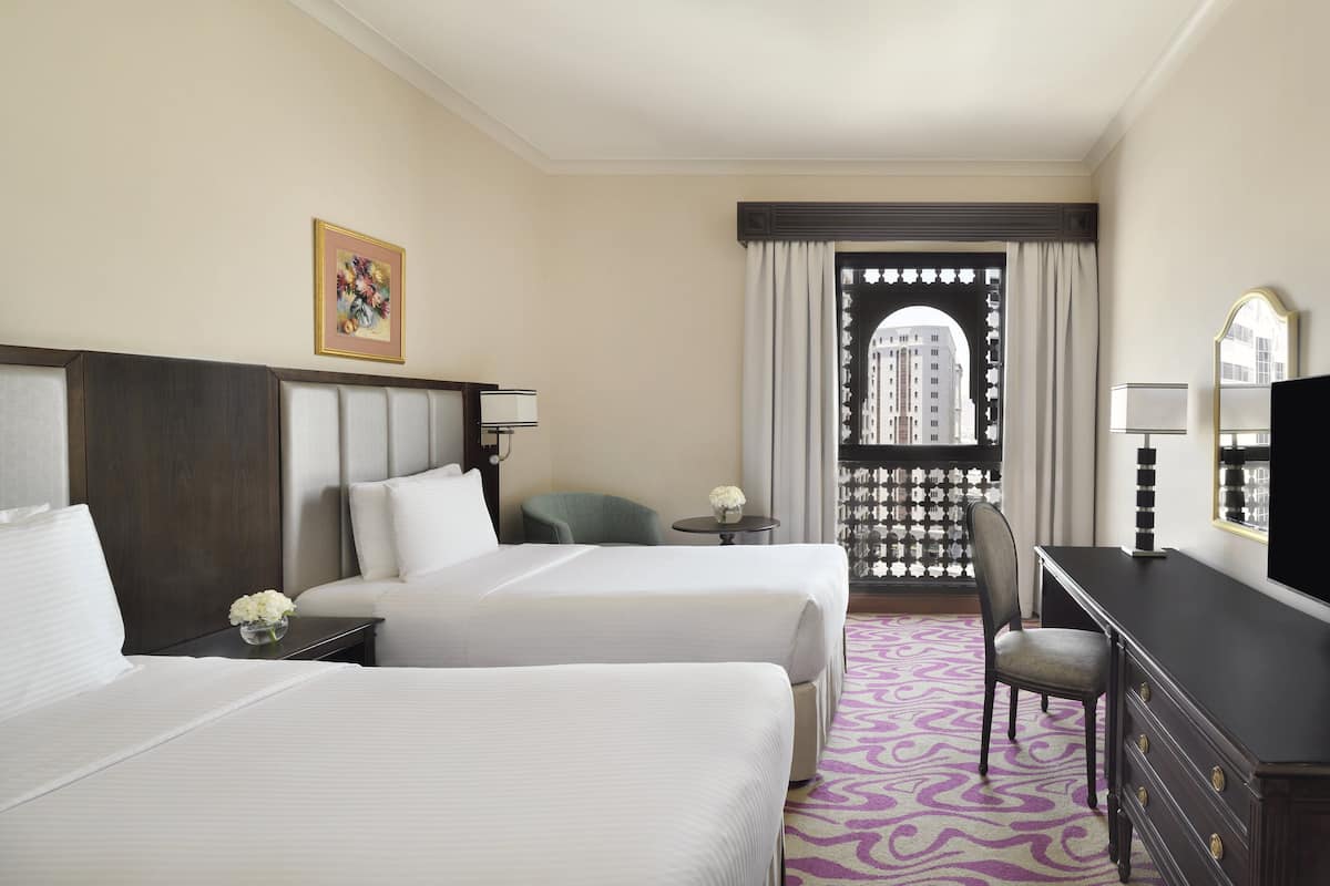 Ein Hotelzimmer mit zwei Betten, einem Schreibtisch, einem Stuhl, einem kleinen Tisch mit einer Vase und einem Fenster mit Vorhängen.