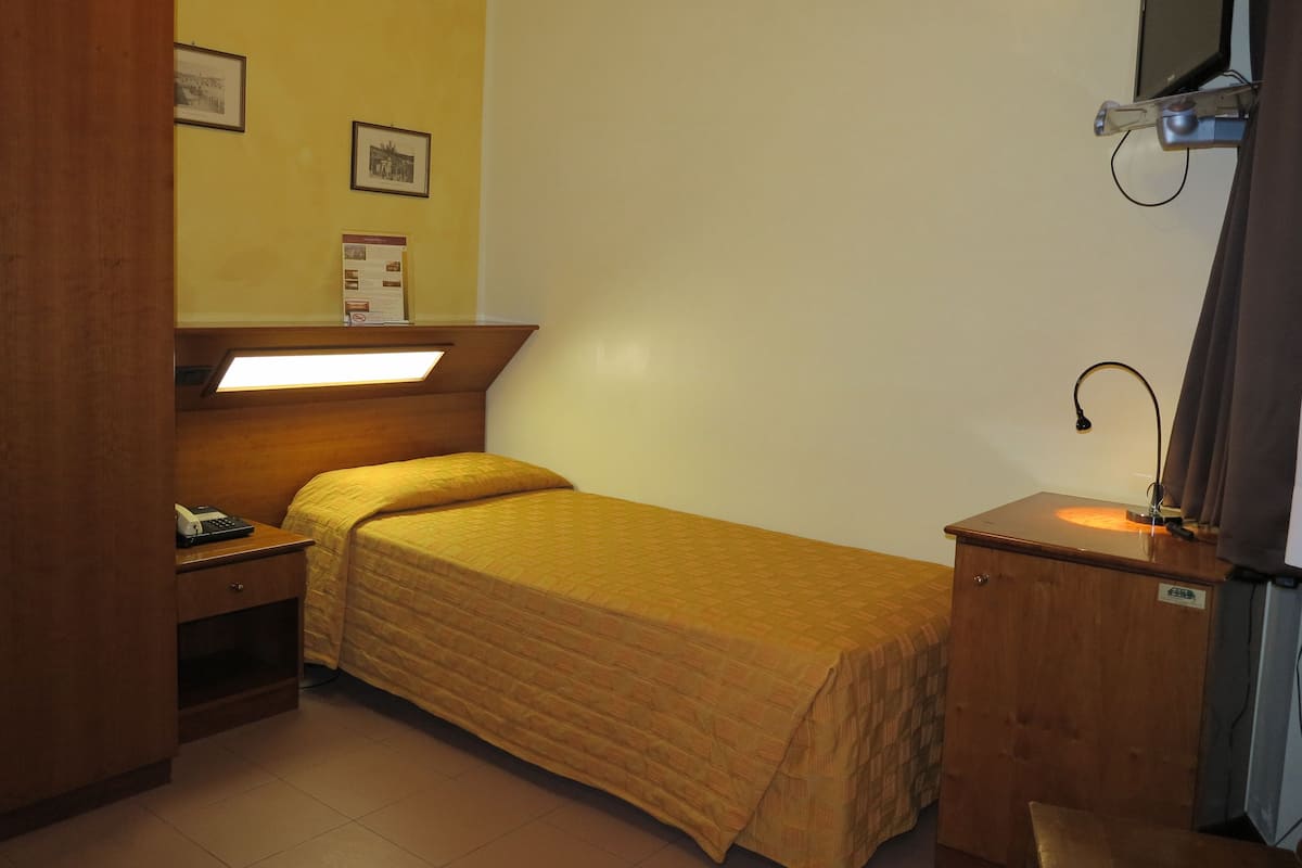 Una habitación pequeña con cama individual, cabecera de madera, mesita de noche con lámpara, escritorio con teléfono y televisor de pantalla plana montado en la pared.