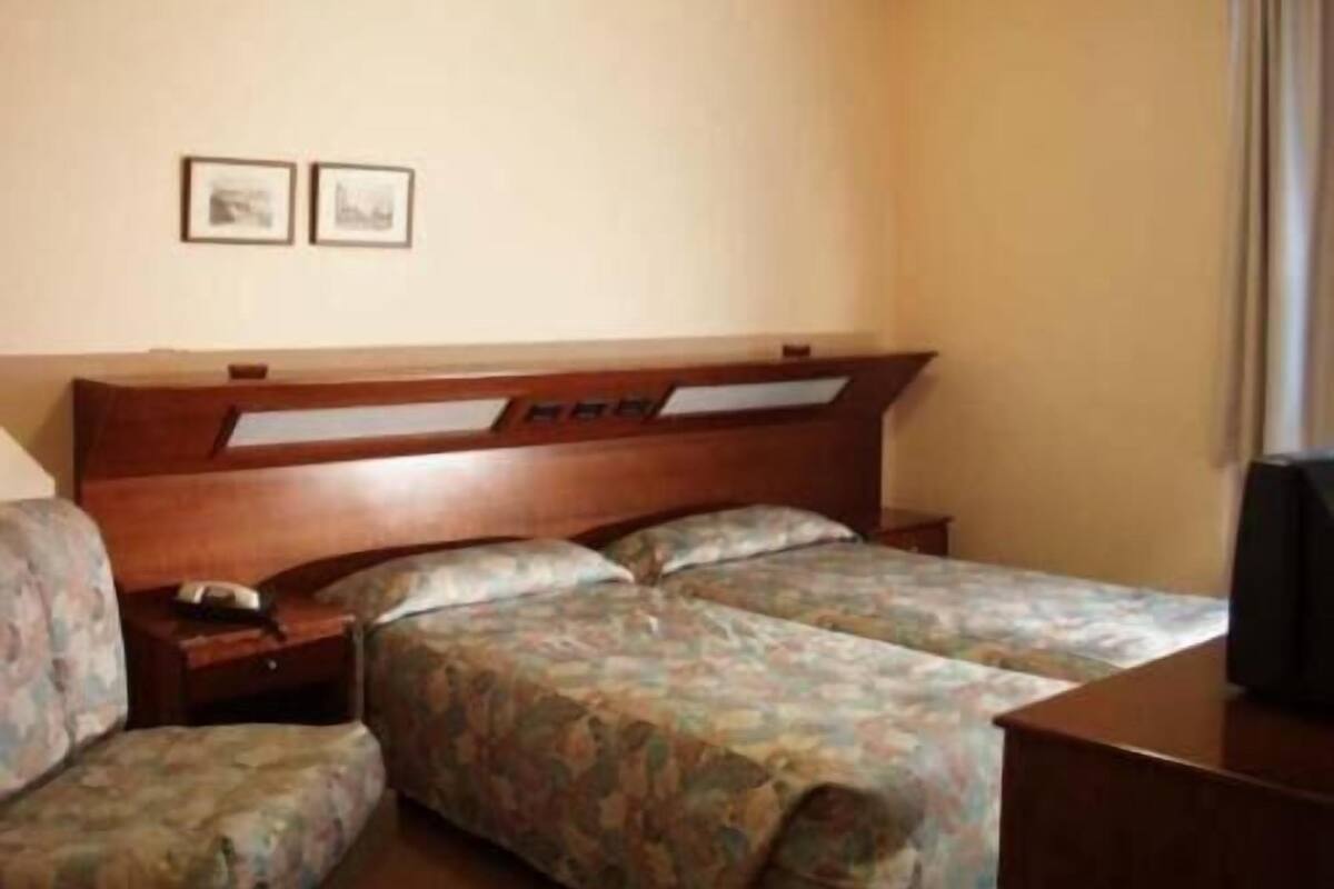 Una habitación de hotel con cabecera de madera, dos camas con ropa de cama floral, una mesita de noche con lámpara y un televisor sobre un mueble.