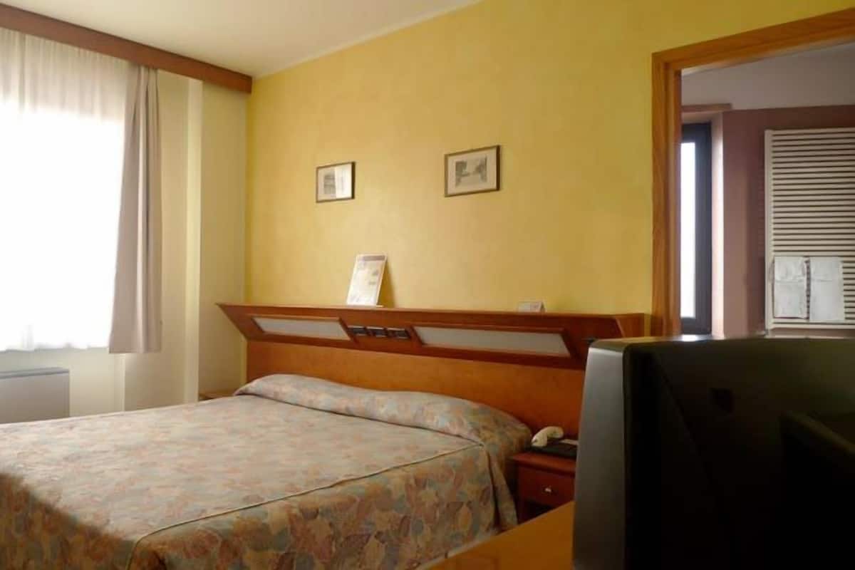 Una habitación de hotel con cama, televisión, mesita de noche y ventana con cortinas.