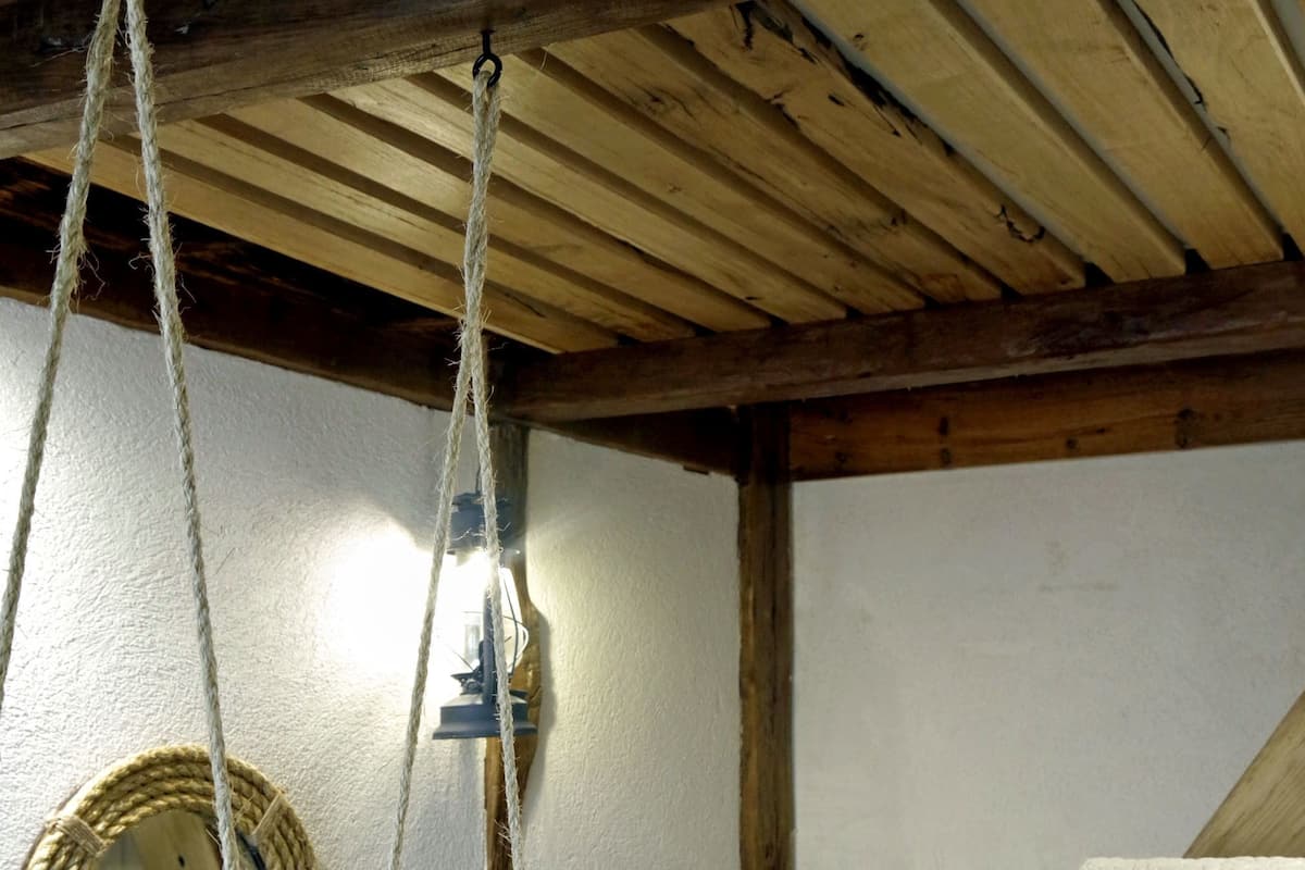 A bedroom with a wooden ceiling, a hanging rope light fixture, a round mirror, and a bed with a blue quilt.