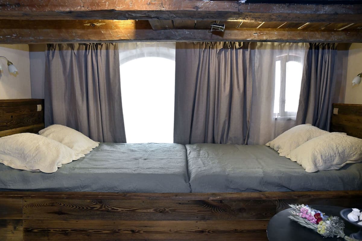 A wooden bed with grey bedding and white pillows, a wooden headboard, and a window with curtains.