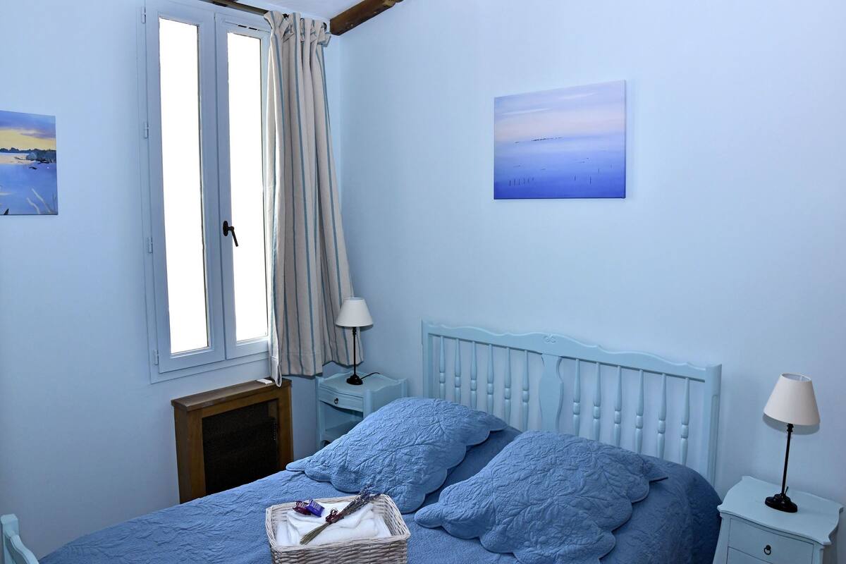 A bedroom with a bed, two bedside tables, a window with curtains, and two wall-mounted paintings.