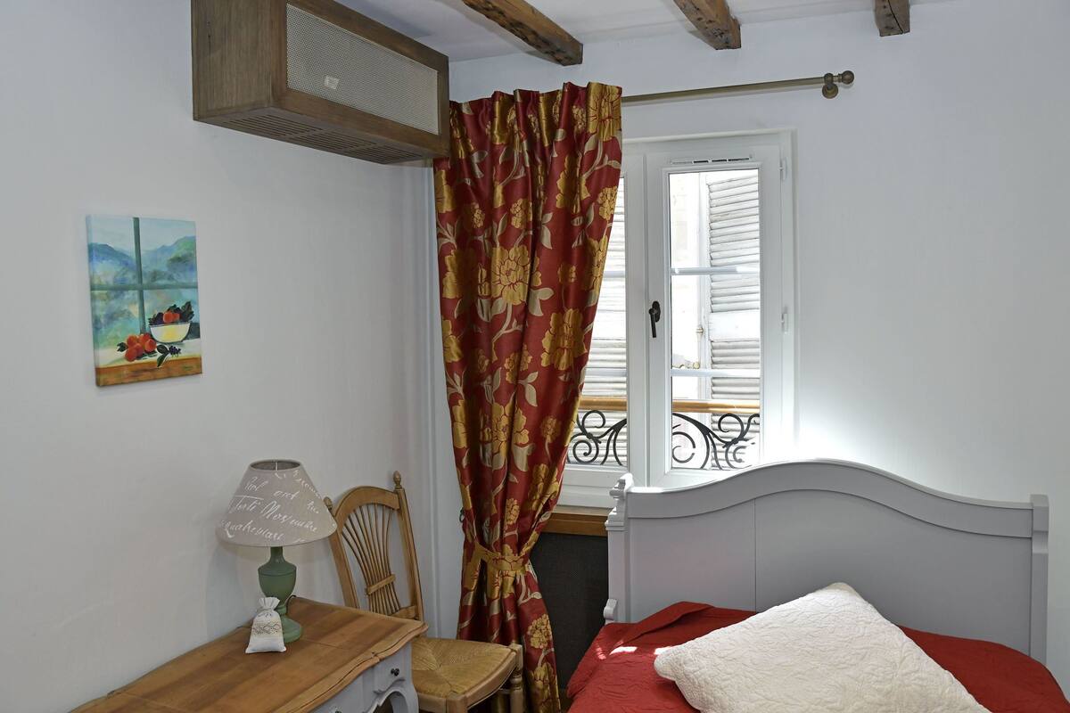 A bedroom with a bed, a chair, a desk with a lamp, a window with blinds, and a painting on the wall.