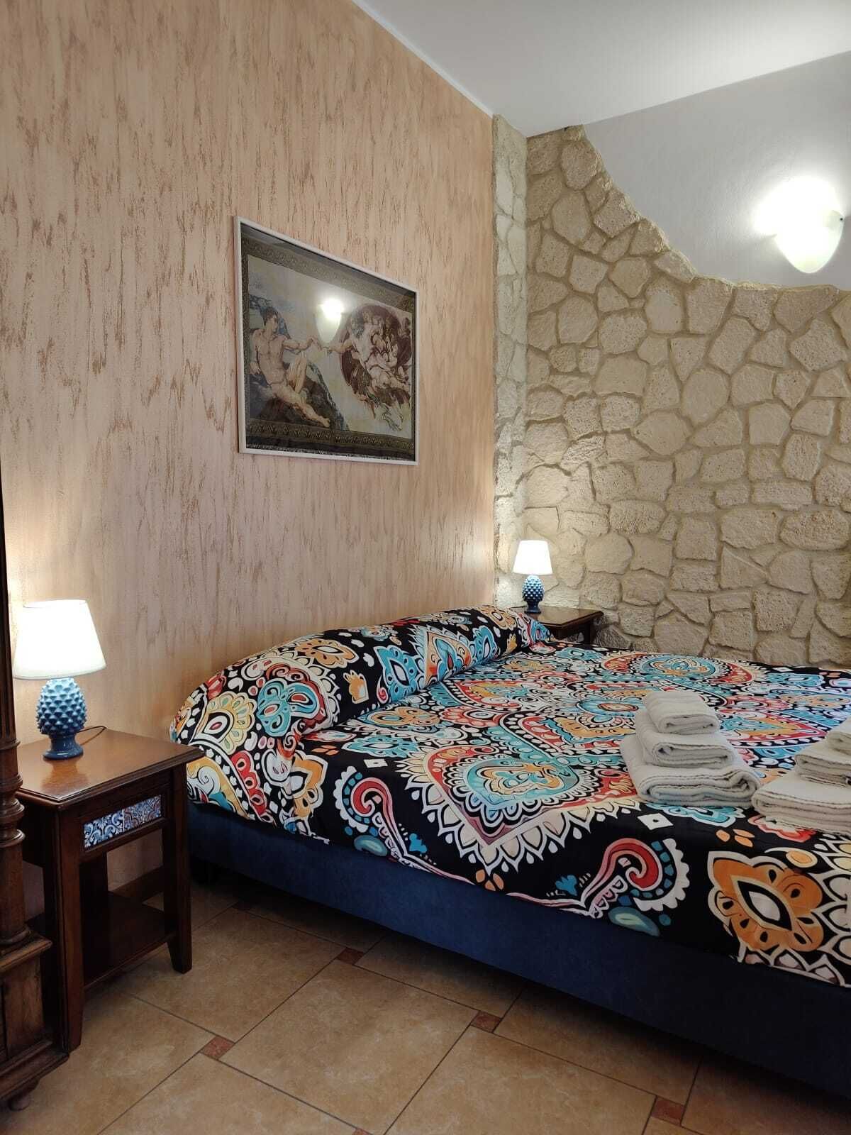 Un dormitorio con cama, mesita de noche, lámpara, un cuadro y pared de piedra.