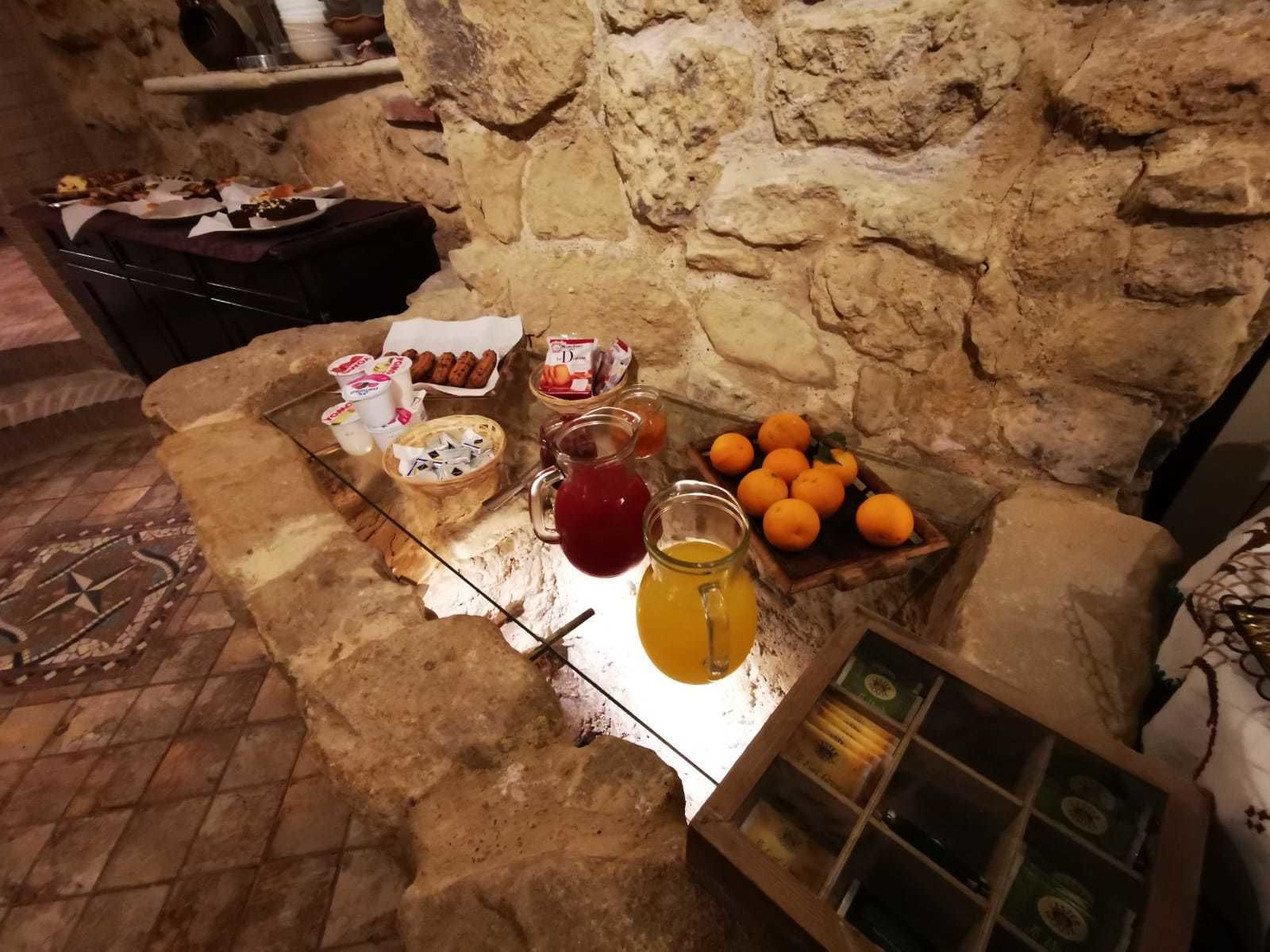 Paredes de pedra com uma mesa de madeira rústica exibindo alimentos como pão, doces e laranjas.