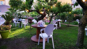 An outdoor seating area with round tables and white chairs, surrounded by greenery and trees. - B&B Proserpina (Enna)