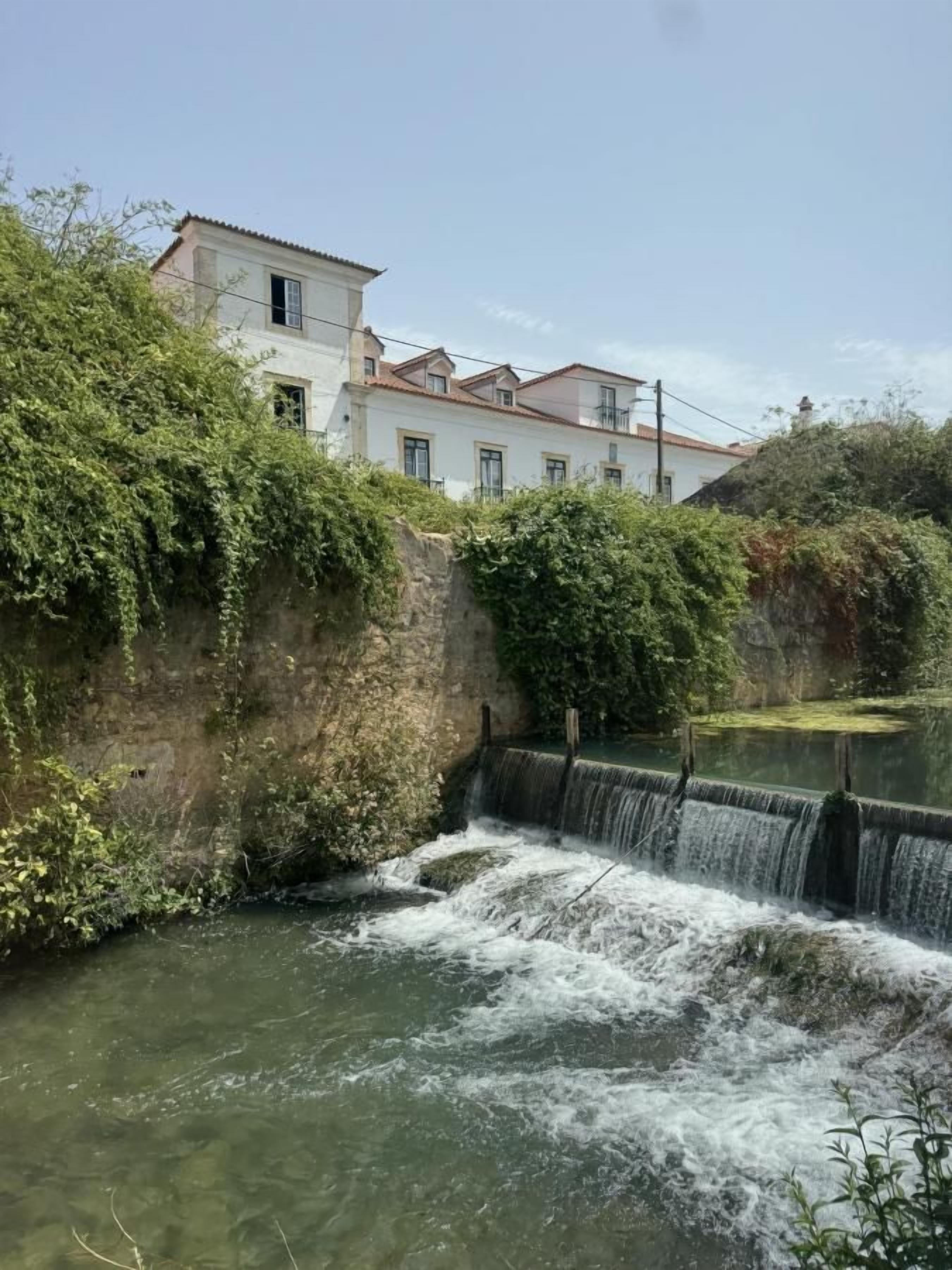 Un bâtiment blanc surmonté d’un toit rouge, entouré de verdure et d’une petite cascade.