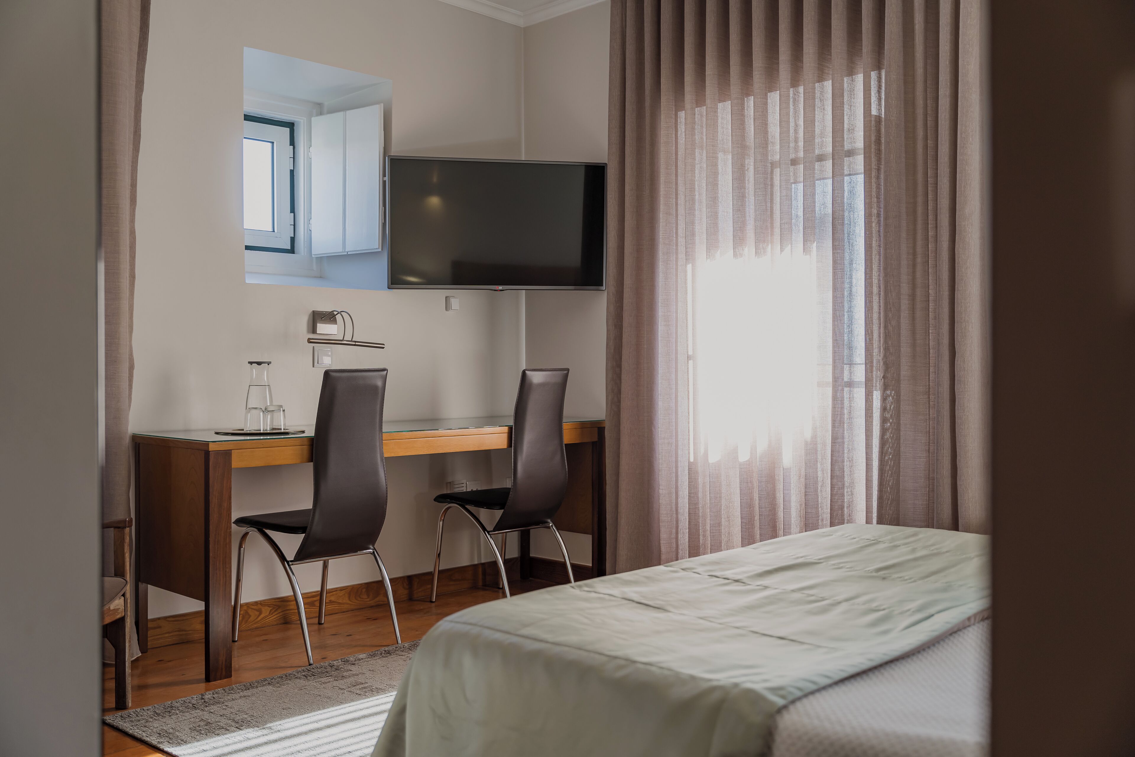 Habitación de hotel con cama, escritorio con dos sillas, televisor de pantalla plana y ventana con cortinas.